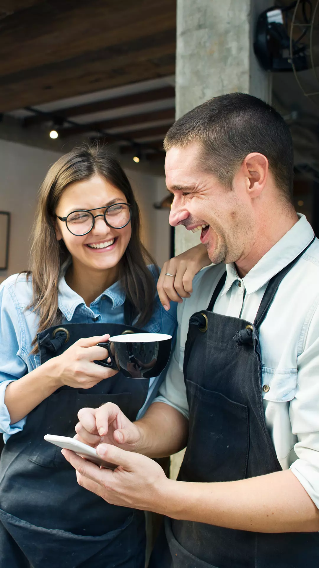 Dois funcionários em um ambiente de trabalho sorrindo, enquanto um toma café o outro mexe ao celular.
